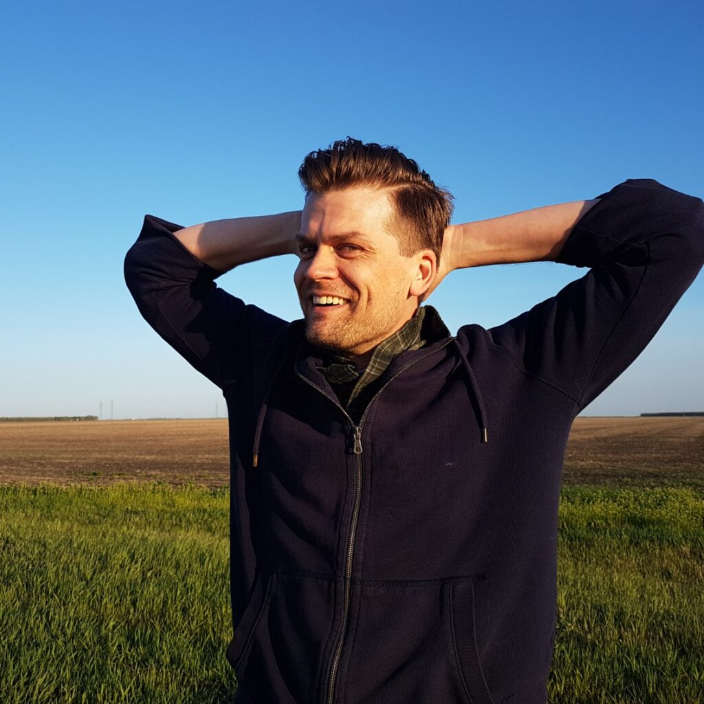 A photo of David smiling, behind him is a blue sky and a field